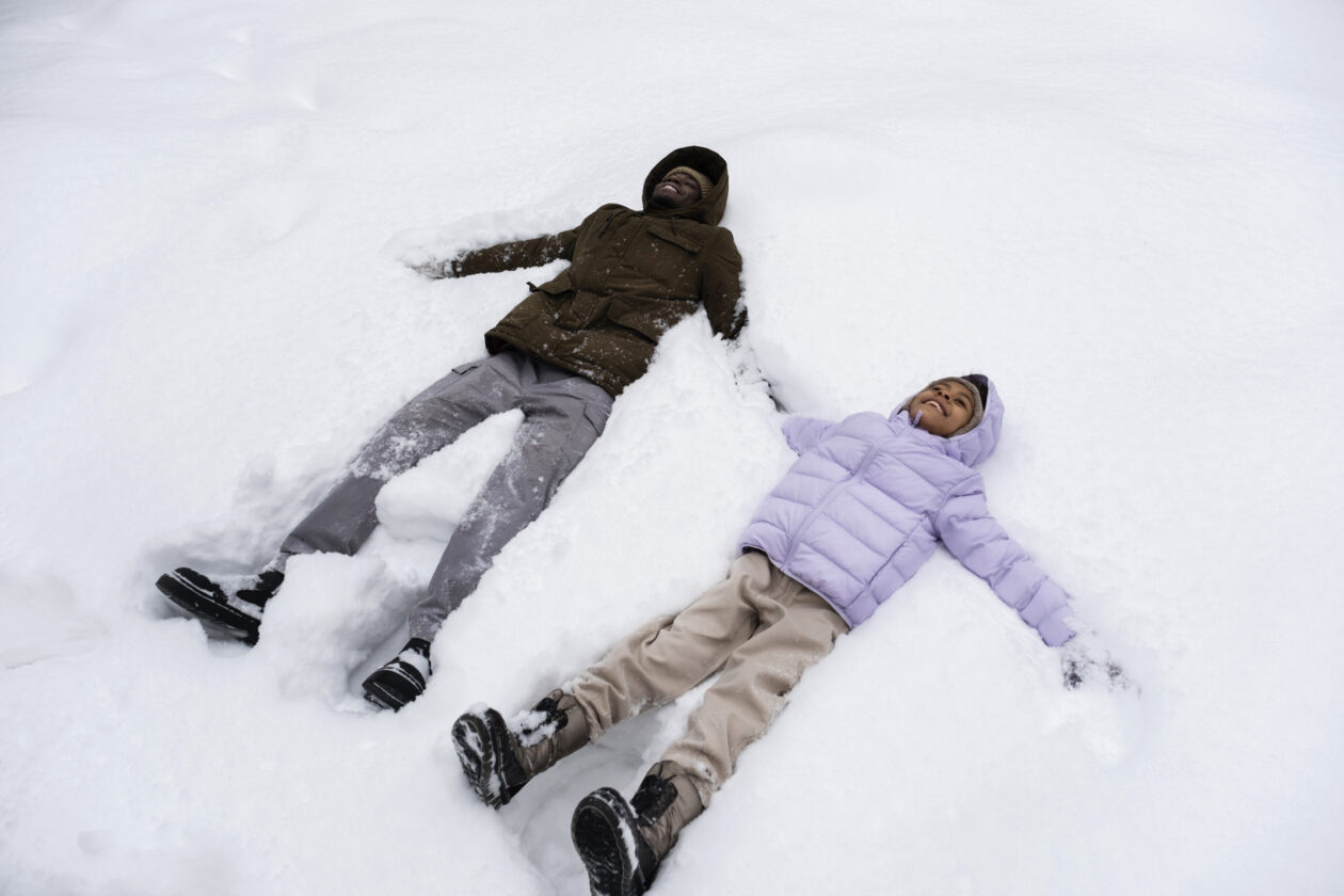 Pai e filha brincando na neve - Viajar com Crianças no Inverno - young-girl-making-snow-angels-with-her-father-winter Viagem para a neve com filhos crescidos: um convite à conexão na transição para a adolescência